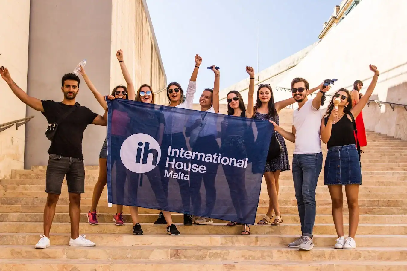 Séjour linguistique à St Julian's : groupe de jeunes étudiants posant avec le drapeau d'International House (IH) Malta, partenaire de Sherwood School, école de langues à La Baule (éligible CPF). Séjour linguistique à St Julian's : groupe de jeunes étudiants posant avec le drapeau d'International House (IH) Malta, partenaire de Sherwood School, école de langues à La Baule (éligible CPF).